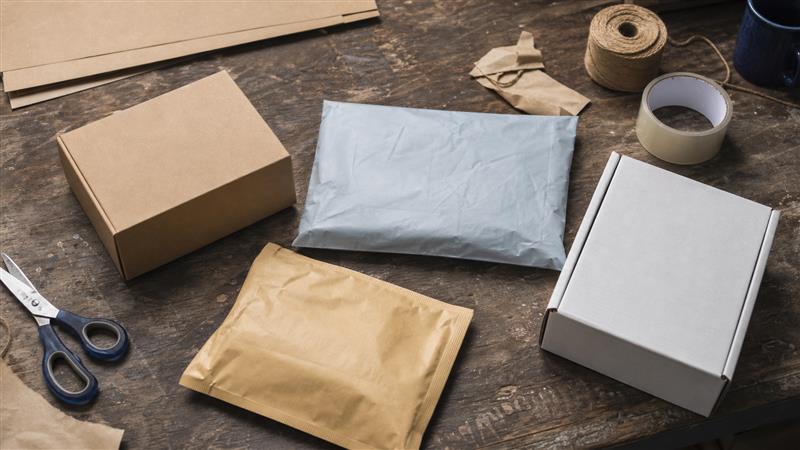 Plain kraft box, gray paper mailer, padded mailer and white corrugated carton on a worn packing table
