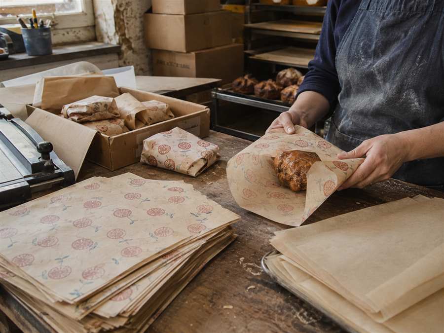 Bakery worker folding custom printed wax paper around pastries at a worn packing table